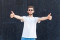 European bearded man in white T-shirt, glasses and jeans showing his tongue and thumbs up close-up in an open area Royalty Free Stock Photo