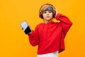 European blond boy in a red hoodie listens to music in red headphones and holds smartphone with mockup on a orange Royalty Free Stock Photo