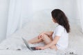 European brunette girl in a white t-shirt, sits on a bed and uses a laptop. Royalty Free Stock Photo