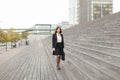 European businesswoman standing on stairs with bag and high buildings in background.