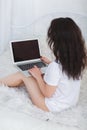 A girl with long dark hair is in a white t-shirt, sits on a bed and uses a modern laptop. Royalty Free Stock Photo