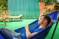 European guy dressed in a blue T-shirt and denim shorts is lying and resting in a blue hammock Royalty Free Stock Photo