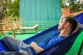 European guy dressed in a blue T-shirt and denim shorts is lying and resting in a blue hammock Royalty Free Stock Photo