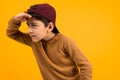 European handsome guy teenager in a brown sweater and baseball cap is looking for a target on a yellow background