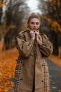 European modern young woman blonde in a stylish coat stands in the park on the road on the background of trees with golden foliage