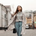 European pretty young brown-haired woman in an elegant trench coat in a knitted vintage sweater in a leather bag in trendy jeans Royalty Free Stock Photo