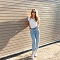 European pretty young hipster woman in stylish white T-shirt in vintage jeans in trendy sunglasses is standing