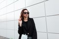 European pretty young hipster woman in trendy sunglasses in a vintage coat in white jeans with a bag stands Royalty Free Stock Photo