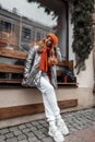 European pretty young woman in fashionable winter clothes sits near a modern building resting on a wooden bench outdoors.