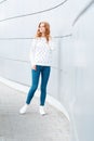 European slim stylish young woman in a vintage sweater in trendy blue jeans in fashionable white sneakers indoors near the wall.