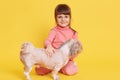 European small dark haired girl pets dog while sitting on floor isolated over yellow background, kid looking at camera with happy Royalty Free Stock Photo