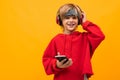 European smiling blond boy in a red hoodie listens to music in red headphones and holds smartphone on a yellow Royalty Free Stock Photo