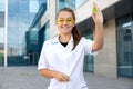 European smiling chemist girl in a white coat and yellow glasses holding a green molecule in hand outside