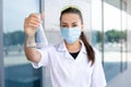 European smiling female biologist in medical gown and protective mask shows Erlenmeyer flask