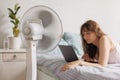 European woman cools down home with help of air conditioner fan, works with laptop in home office