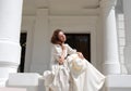 European woman sitting in sunshine and touching hair in vintage dress near palace.