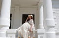 European woman sitting in sunshine and touching hat in vintage dress near palace. Royalty Free Stock Photo