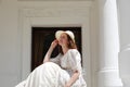 European woman sitting in sunshine and touching hat in vintage dress near palace. Royalty Free Stock Photo