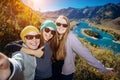 European women in sunglasses laugh doing selfie against mountain background. Young female hikers pose against amazing small island