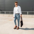 European young man hipster in a white shirt in blue fashionable jeans in dark sunglasses with a black bag posing in the street on Royalty Free Stock Photo