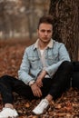 European young man in a stylish denim jacket in trendy military pants in white sneakers with a backpack is resting near a tree in Royalty Free Stock Photo