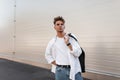 European young man with a stylish hairstyle in a trendy white shirt in vintage jeans is standing near a modern white building Royalty Free Stock Photo