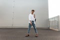 European young man in trendy white and denim clothes in fashionable red sandals with a vintage bag walks on the city. Modern