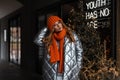 European young red-haired woman in a stylish knitted hat with a trendy orange scarf in a youth fashionable silver jacket posing