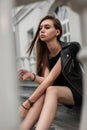 European young woman model in an elegant short black dress in a stylish black leather jacket sits on a stone staircase near a Royalty Free Stock Photo