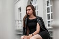 European young woman model in an elegant short black dress in a stylish black leather jacket sits on a vintage staircase near a Royalty Free Stock Photo