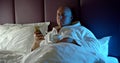 In the evening in the bed a bald adult man lies under a white blanket with a cup of tea and watches the news