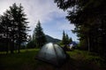 Evening camping at mountains. Friends are sitting near tents in which light is on between fir trees under evening sky Royalty Free Stock Photo