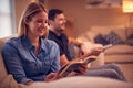 Evening Shot Of Couple Relaxing On Sofa At Home As Woman Reads Book And Man Watches TV Royalty Free Stock Photo