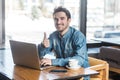 Everything alright! Portrait of handsome satisfied bearded young freelancer in blue jeans shirt are sitting in cafe and working on