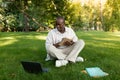 Exam study concept. Black male student guy taking notes while watching online lesson on laptop, sitting on lawn in park Royalty Free Stock Photo