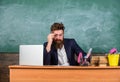 Examiner full of doubts sit at table chalkboard background. School exam concept. Examiner bearded teacher with