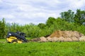 Excavator digging pit on grassy field with garden on background