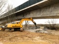 An excavator running a concrete demolition with a hydraulic breaker under a bridge under construction