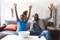 Excited african american father and son sitting on sofa, watching match on tv and cheering