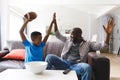 Excited african american father and son sitting on sofa, watching match on tv and cheering