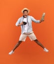 Excited black man with pineapple and professional camera jumping, straw hat flying above his head on orange background