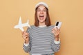 Excited amazed woman wearing striped shirt and baseball cap holding white paper plane and using smartphone booking tickets to Royalty Free Stock Photo