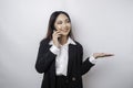 Excited Asian businesswoman wearing black suit pointing at the copy space beside her while talking on her phone, isolated by white Royalty Free Stock Photo