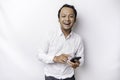 Excited Asian man wearing white shirt smiling while holding his phone, isolated by white background Royalty Free Stock Photo