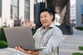 Excited asian mature businessman having video call on laptop, waving hand to webcamera, sitting near office building