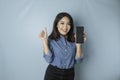 Excited Asian woman wearing a blue shirt gives thumbs up hand gesture of approval while holding her smartphone, isolated by blue Royalty Free Stock Photo