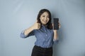Excited Asian woman wearing a blue shirt gives thumbs up hand gesture of approval while holding her smartphone, isolated by blue Royalty Free Stock Photo