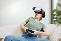 Excited attractive woman gaming with virtual reality headset or goggles and sitting on the couch, holding gamepad controller and