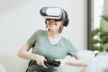 Excited attractive woman gaming with virtual reality headset or goggles and sitting on the couch, holding gamepad controller and