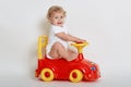 Excited baby toy sitting on red and yellow tolocar and looking at camera with satisfied facial expression, wearing bodysuit, Royalty Free Stock Photo